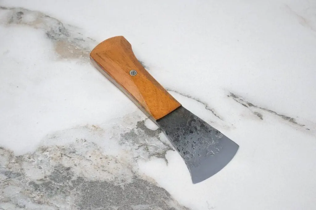 The March Madness 3 Piece Essentials set, featuring a hand axe with a wooden handle and metal blade, elegantly rests on a marble surface as part of your outdoor essentials.