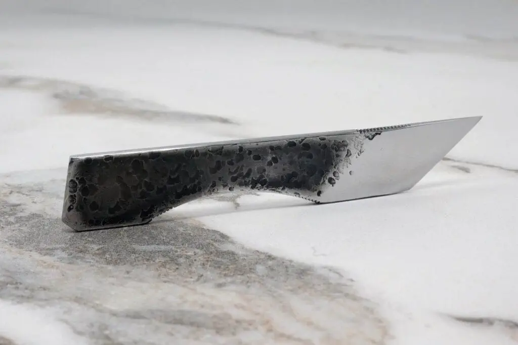 A modern knife with a sleek, angular design and a textured black handle rests on a marble surface.