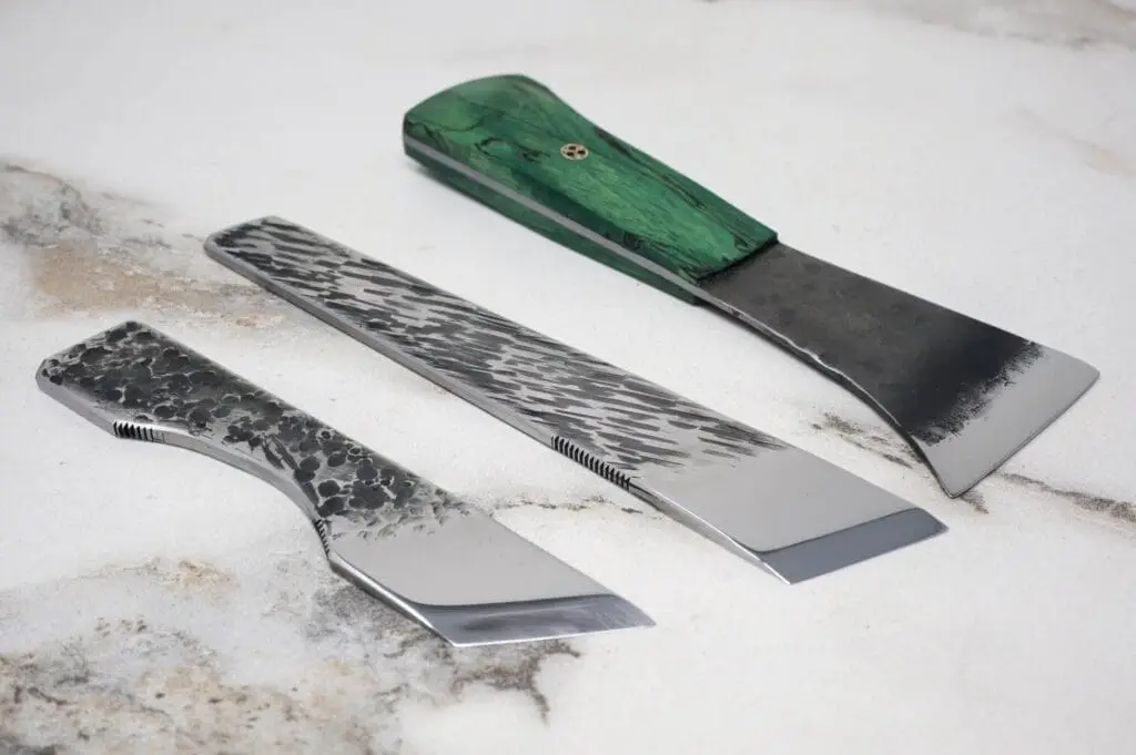 Three metal straight razors with uniquely textured blades and handles are displayed on a marbled surface.