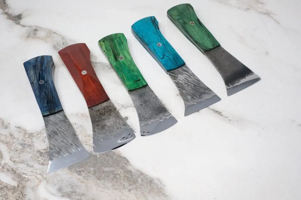 Five Forged American Signature Knives with colored wooden handles in blue, red, and green are elegantly arranged on a marbled surface.